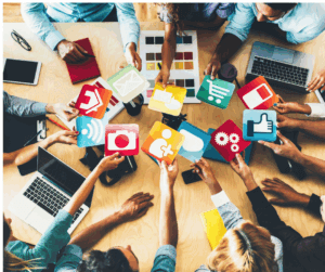 A diverse group of people sit around a wooden table, collaborating and holding up colorful icons representing social media, communication, shopping, technology, and networking. The table is scattered with laptops, notebooks, and color swatches, symbolizing teamwork, creativity, and digital connection.