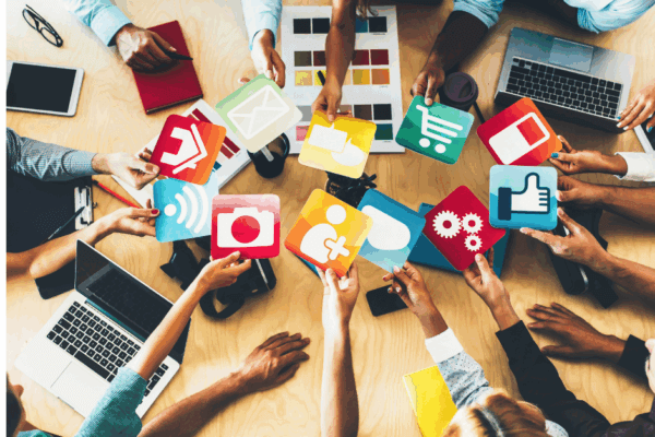 A diverse group of people sit around a wooden table, collaborating and holding up colorful icons representing social media, communication, shopping, technology, and networking. The table is scattered with laptops, notebooks, and color swatches, symbolizing teamwork, creativity, and digital connection.